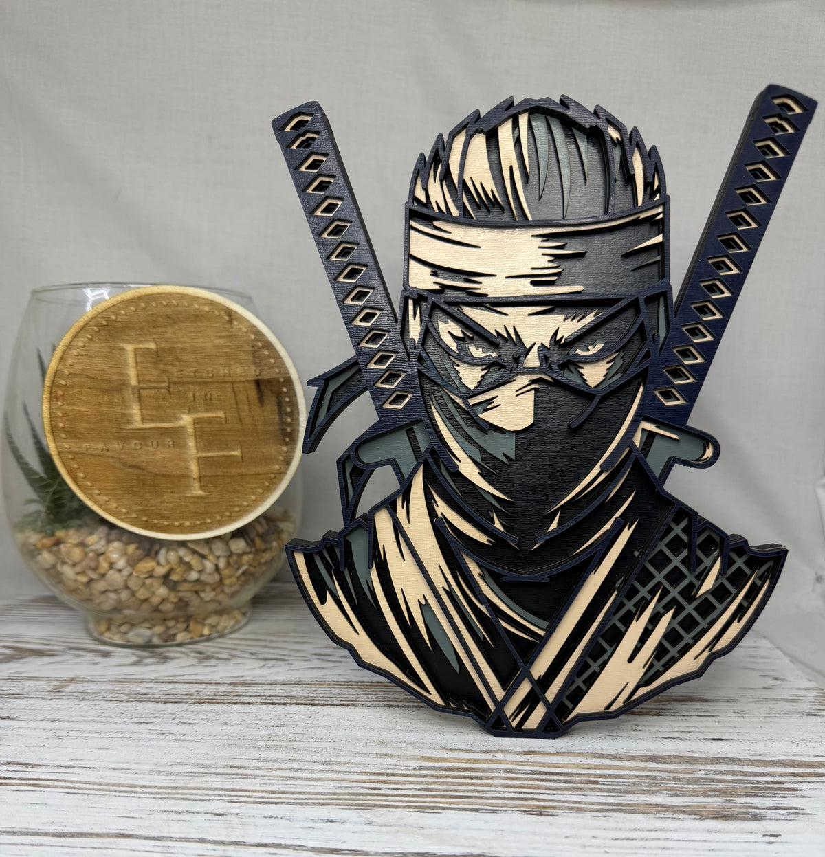 A laser-cut wooden decorative figurine of a ninja warrior with two crossed swords, made from colored wood from Etched in Favour.