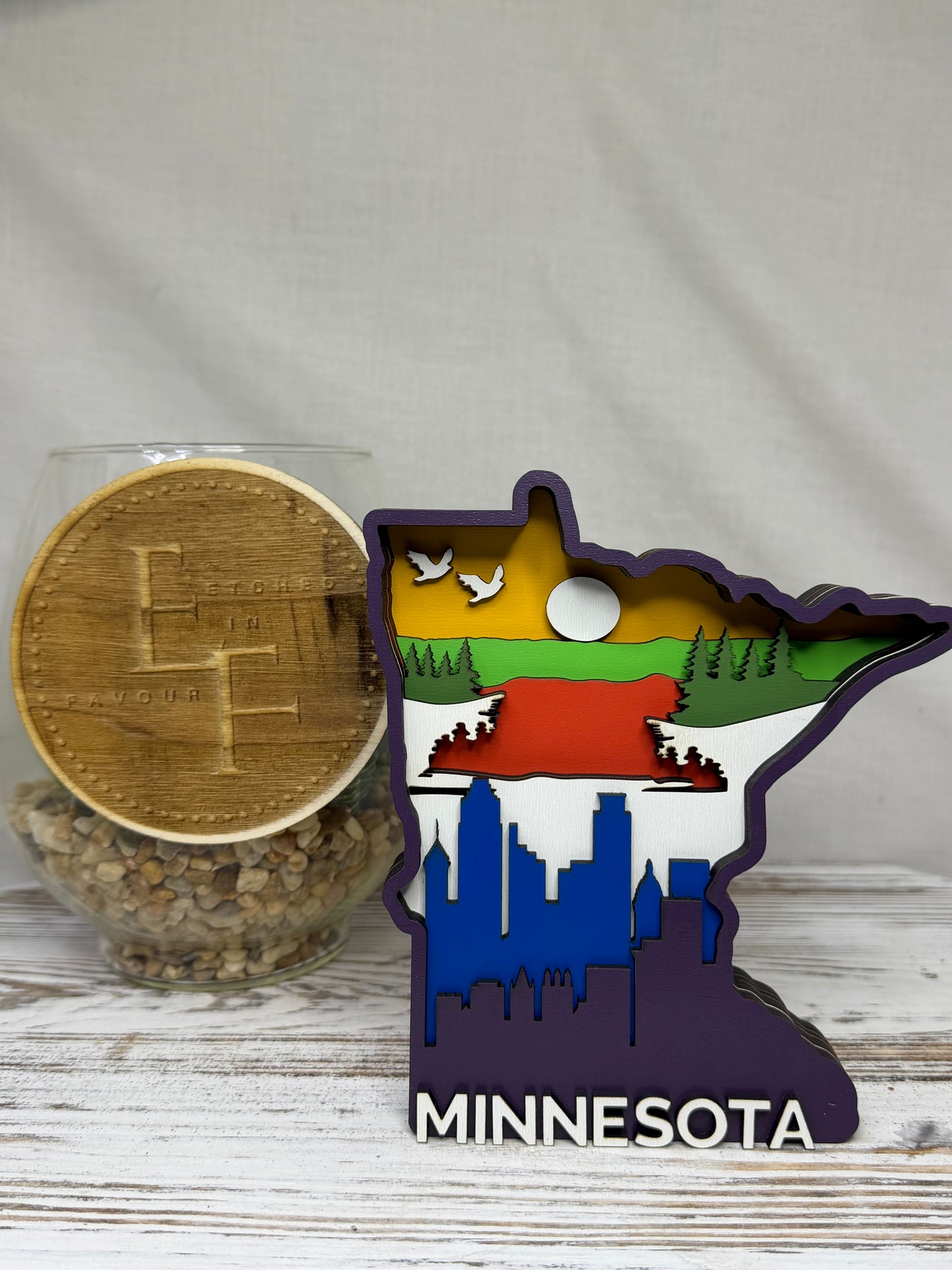 A laser cut multilayered and multicolor wooden Minnesota-shaped decor piece featuring a city skyline, forestry, and birds flying with the word Minnesota at the bottom, from Etched in Favour