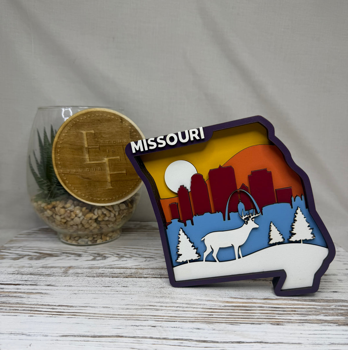 A laser cut multilayered and multicolor wooden Missouri-shaped decor piece featuring a layered winter/sunset cityscape with the word MISSOURI on top from Etched in Favour.