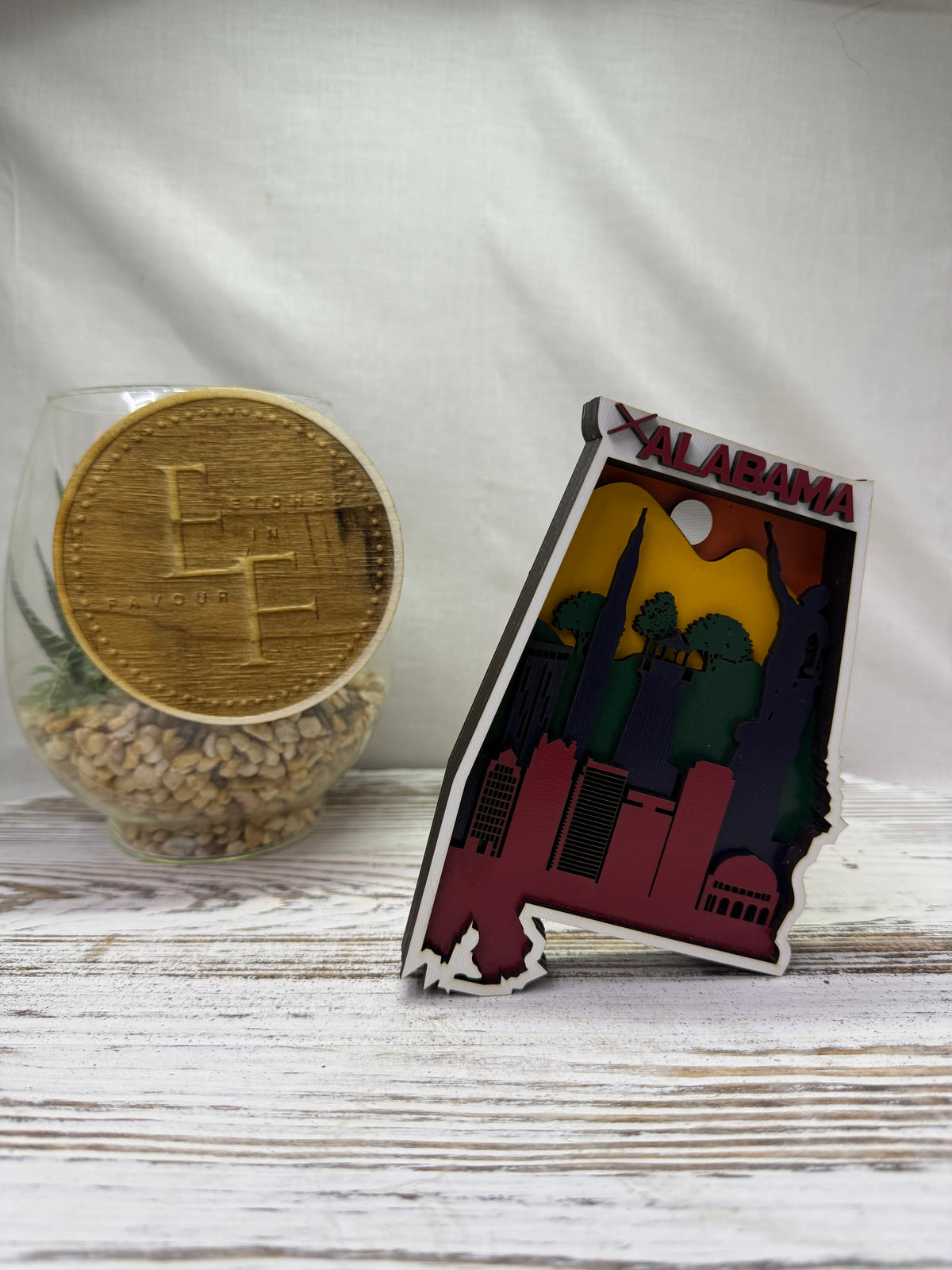A laser cut multilayered and multicolor wooden Alabama state-shaped decor piece featuring a colorful skyline and the word 'ALABAMA' in red, from Etched in Favour.