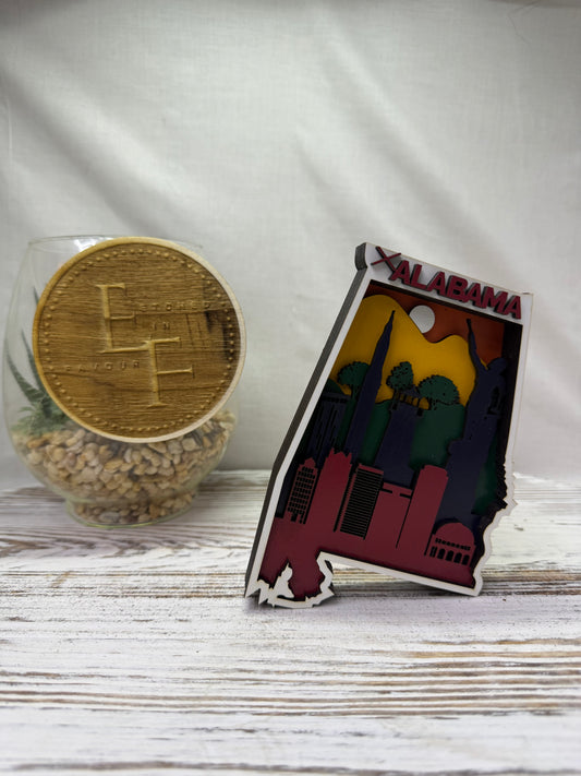 A laser cut multilayered and multicolor wooden Alabama state-shaped decor piece featuring a colorful skyline and the word 'ALABAMA' in red, from Etched in Favour.