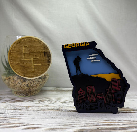 A laser cut multilayered and multicolor wooden Georgia state-shaped decor piece featuring a skyline design, with a hiker looking over a mountain cliff and the word 'Georgia' in yellow at the top from Etched in Favour.