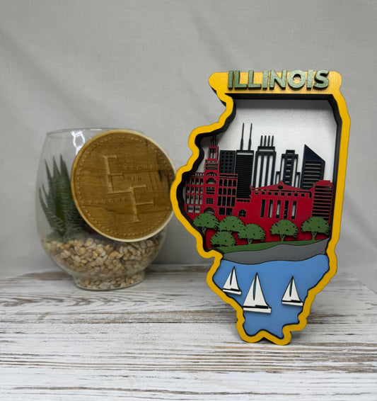 A laser cut multilayered and multicolor wooden Illinois state-shaped decor piece featuring a city skyline, landscape with water and boats, and the word 'Illinois' in green at the top from Etched in Favour.