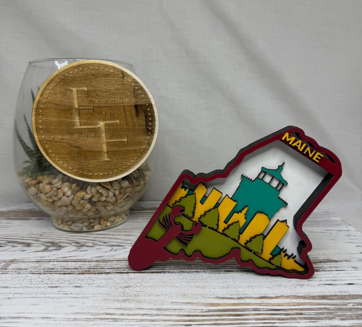 A laser cut multilayered and multicolor wooden Maine state-shaped decor piece featuring a city skyline, landscape and lobster with the word 'Maine' in yellow at the top from Etched in Favour.