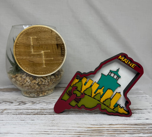 A laser cut multilayered and multicolor wooden Maine state-shaped decor piece featuring a city skyline, landscape and lobster with the word 'Maine' in yellow at the top from Etched in Favour.