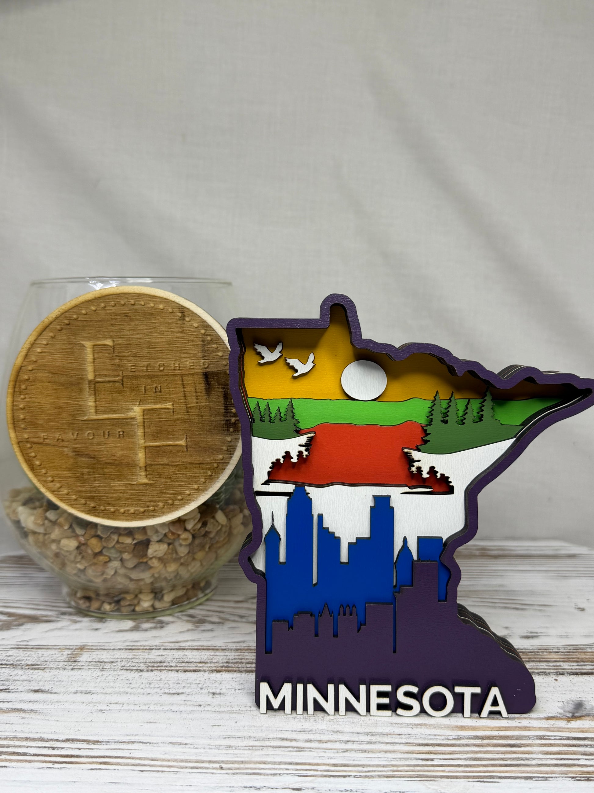 A laser cut multilayered and multicolor wooden Minnesota-shaped decor piece featuring a city skyline, forestry, and birds flying with the word Minnesota at the bottom, from Etched in Favour