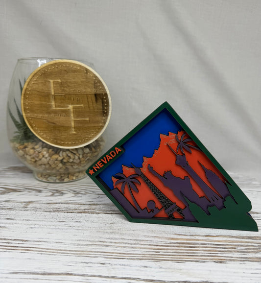 A laser cut multilayered and multicolor wooden Nevada state-shaped decor piece featuring orange mountains, palm trees, and features of the Las Vegas strip with the word 'Nevada' in orange at the top from Etched in Favour.