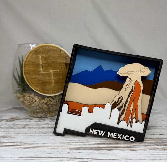 A laser cut multilayered and multicolor wooden New Mexico-themed decor piece featuring a layered landscape design including blue mountains, beige and brown hills, an orange volcanic shape, and a white city silhouette at the bottom with the words 'NEW MEXICO' from Etched in Favour.