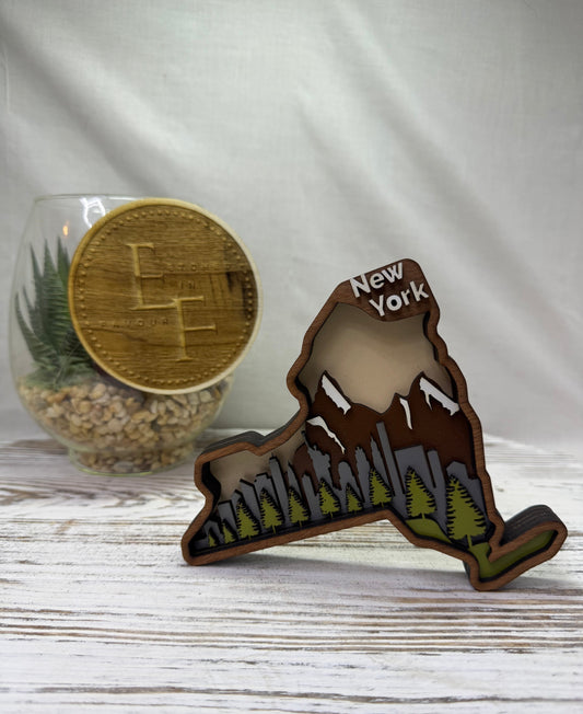 A laser cut multilayered and multicolor wooden New York City skyline-shaped decor piece featuring a brown border, a gray city skyline, green trees, brown snowcapped mountains and the words 'NEW YORK' on the top from Etched in Favour.