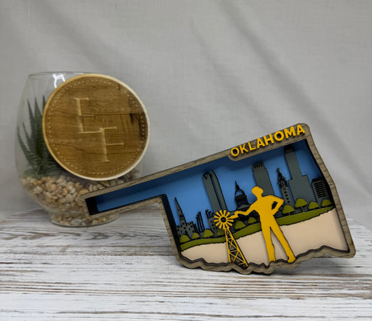 A laser cut multilayered and multicolor wooden Oklahoma-shaped decor featuring a yellow silhouette of a farmer near a windmill, a blue skyline, green hills, and the word 'Oklahoma' in yellow from Etched in Favour