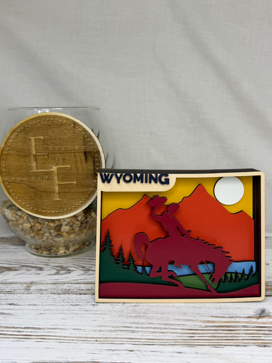 A laser cut multilayered and multicolor wooden Wyoming-themed decor piece featuring a mountain and cowboy silhouette design, with water and forestry and the word Wyoming, from Etched in Favour. 